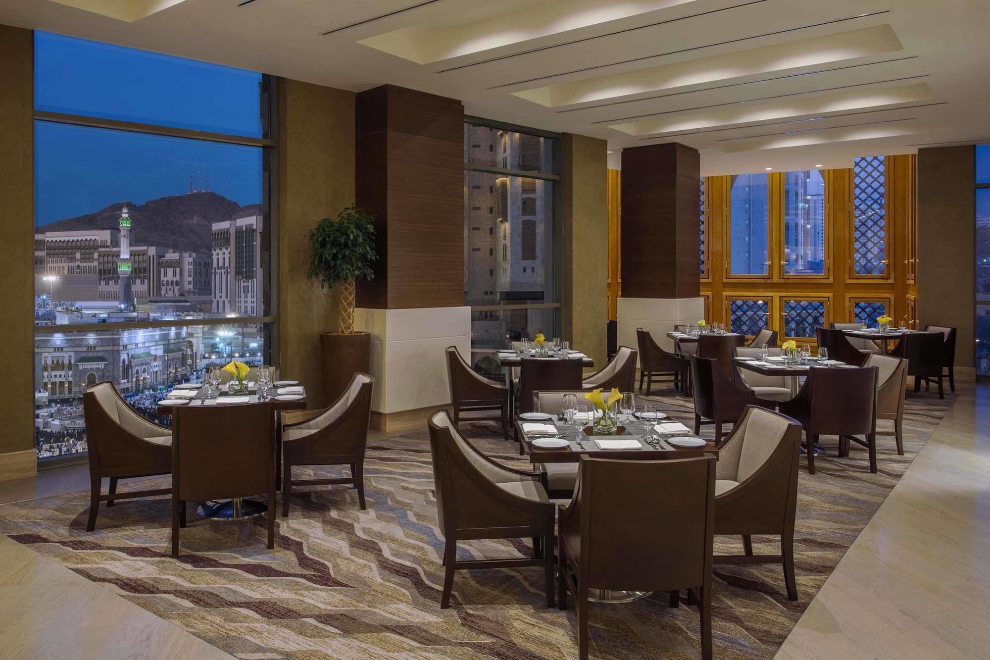 Jabal Omar Hyatt Regency Makkah Hotel Mekka Kültér fotó The photo shows a sophisticated restaurant interior, featuring elegant round tables with high-backed chairs arranged neatly. The space is well-lit with large windows that provide a view of the city skyline at dusk, with mountains in the background. T