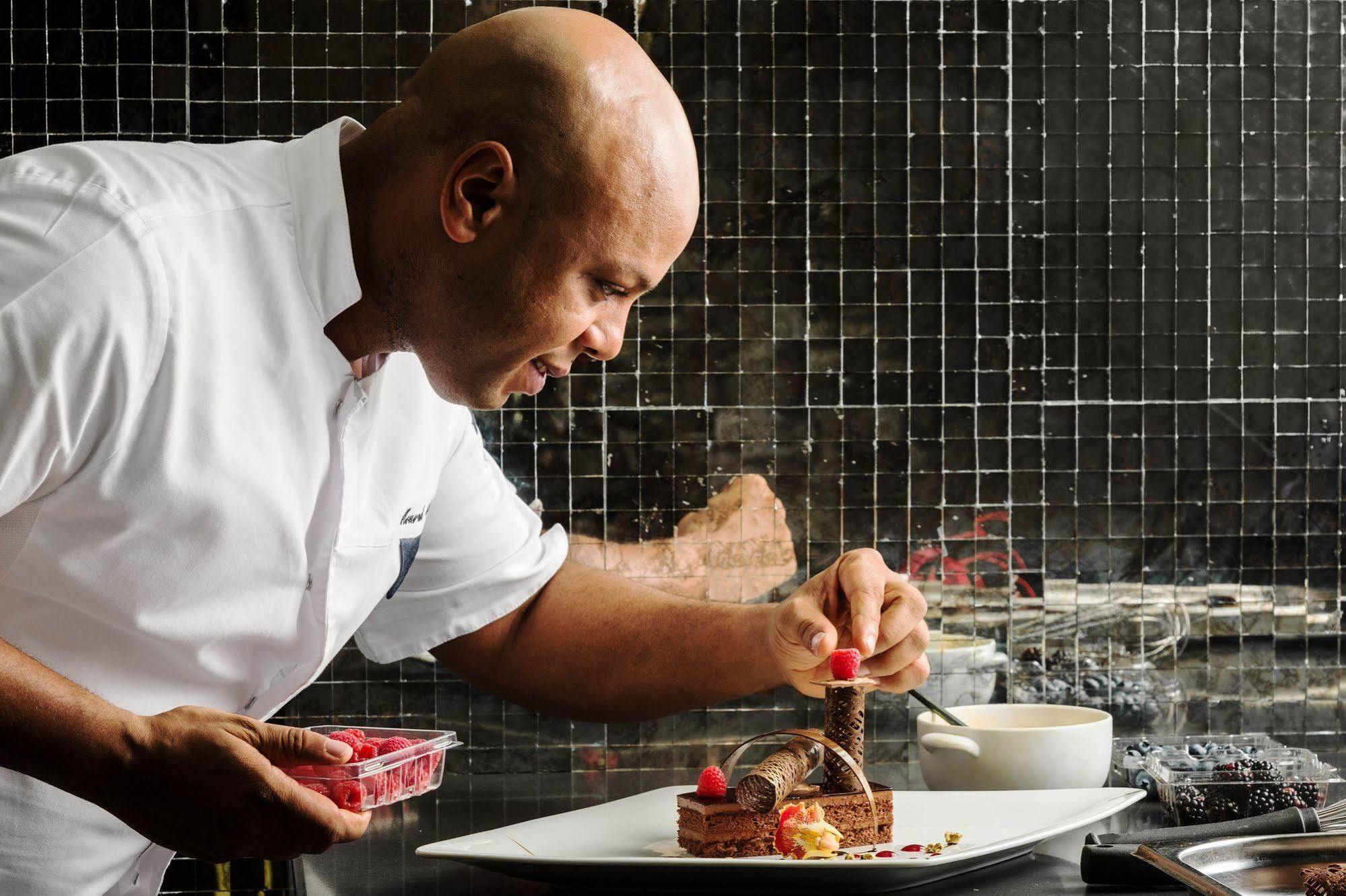 Jabal Omar Hyatt Regency Makkah Hotel Mekka Kültér fotó The photo depicts a chef, who is bald and wearing a white chef's coat, carefully plating a dessert. He is focused on adding a finishing touch, possibly a raspberry, to the dessert on a large white plate. The background features a tiled wall, and ther