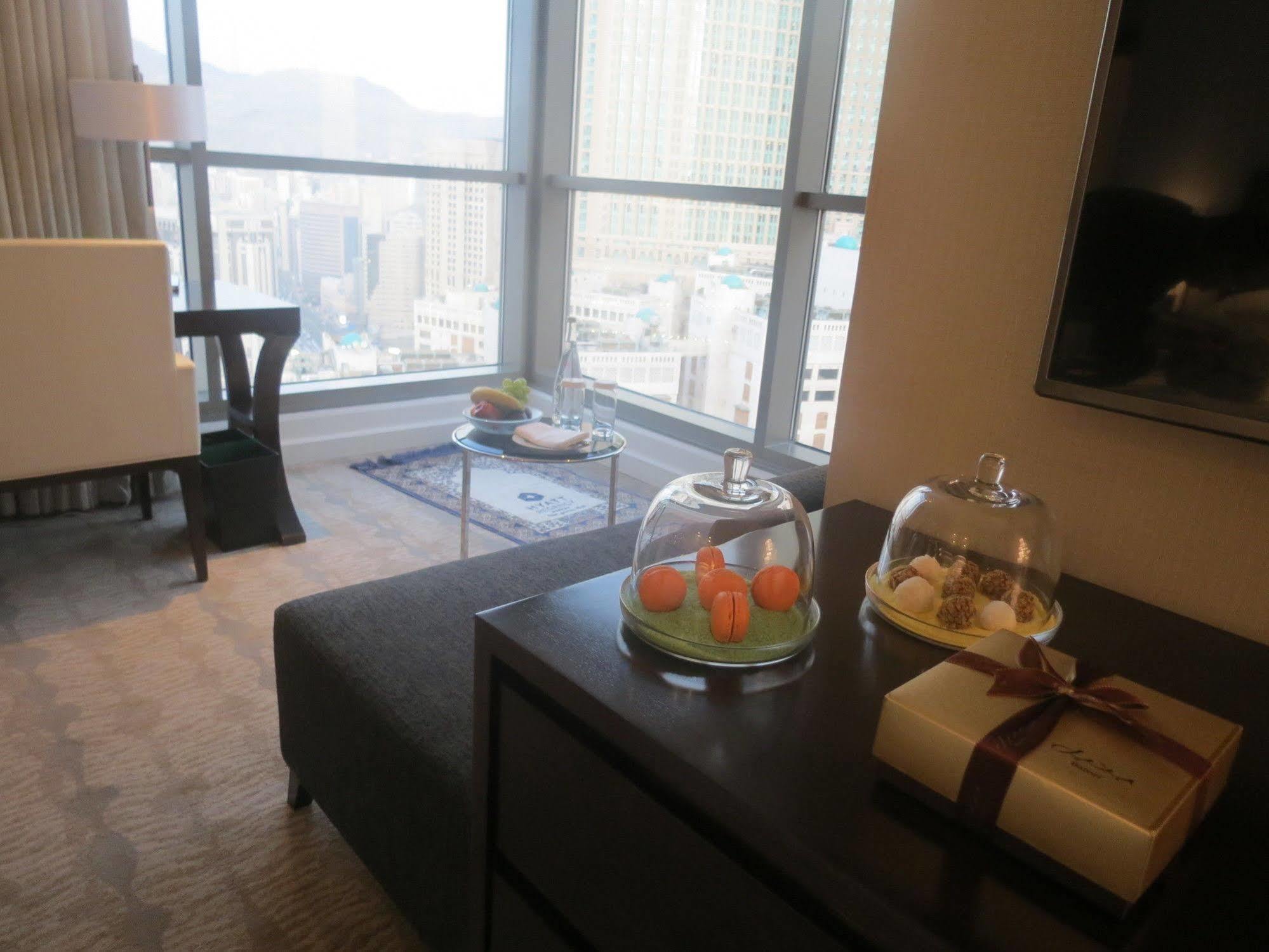 Jabal Omar Hyatt Regency Makkah Hotel Mekka Kültér fotó The photo shows a modern hotel room or a stylish living space with large windows offering a view of the city. In the foreground, there is a dark sofa and a low table with covered trays displaying what appears to be small desserts or delicacies. To th
