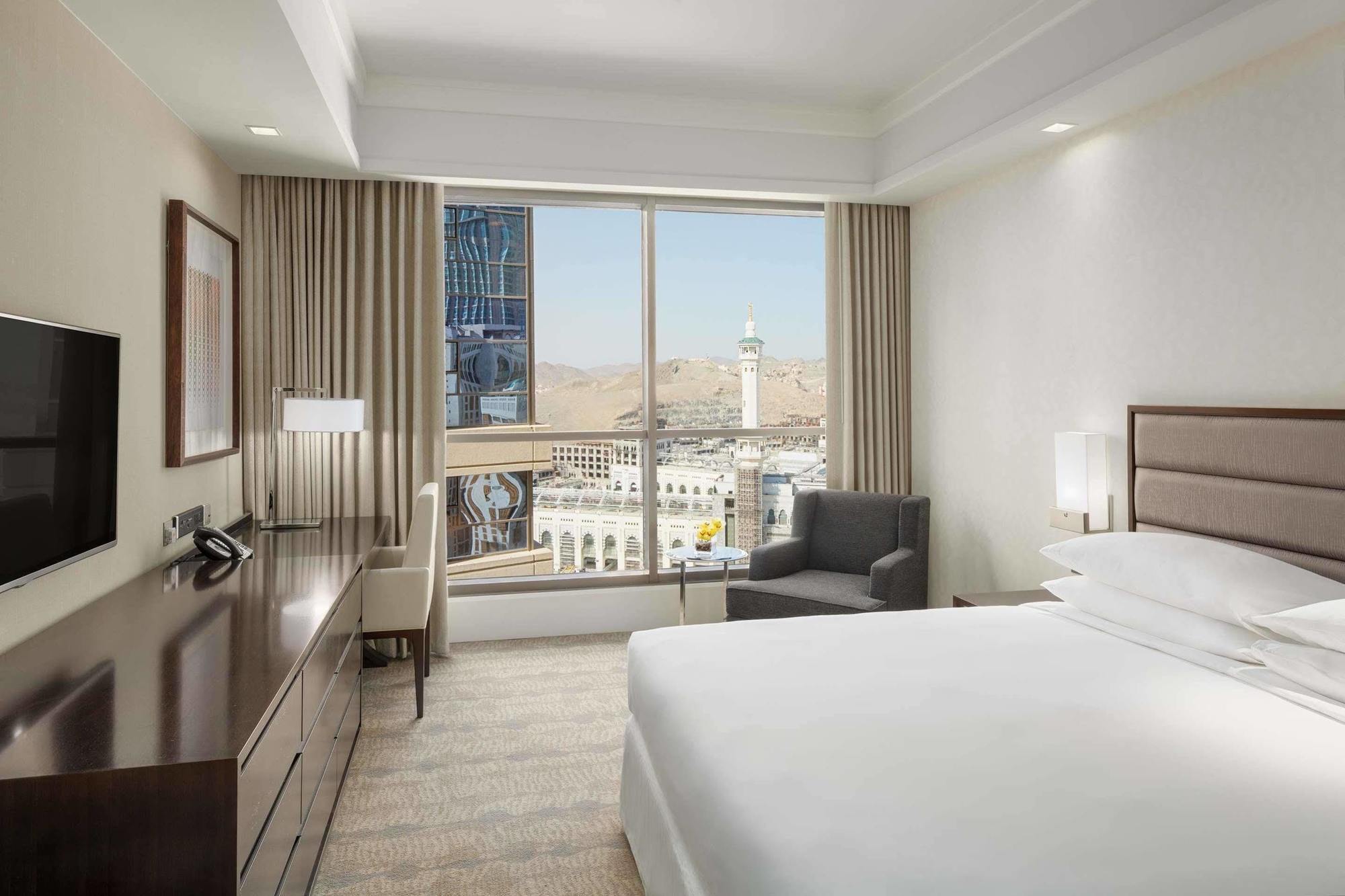 Jabal Omar Hyatt Regency Makkah Hotel Mekka Kültér fotó The photo shows a modern hotel room featuring a large window that provides a view of an outdoor landscape, likely part of a city or landmark. The room includes a bed with white linens, a side table with a lamp, and a small seating area with a chair a
