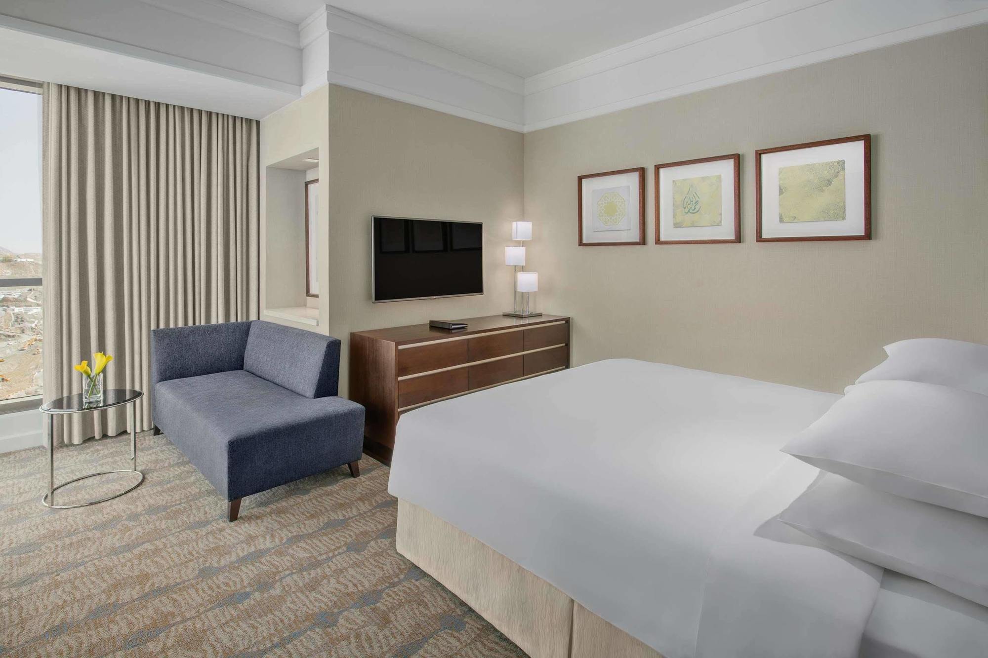 Jabal Omar Hyatt Regency Makkah Hotel Mekka Kültér fotó The photo shows a modern hotel room featuring a neatly made bed with white linens, a small blue couch to the side, and a wooden dresser with a television mounted on the wall above it. There are two framed pieces of art on the wall near the bed, and t