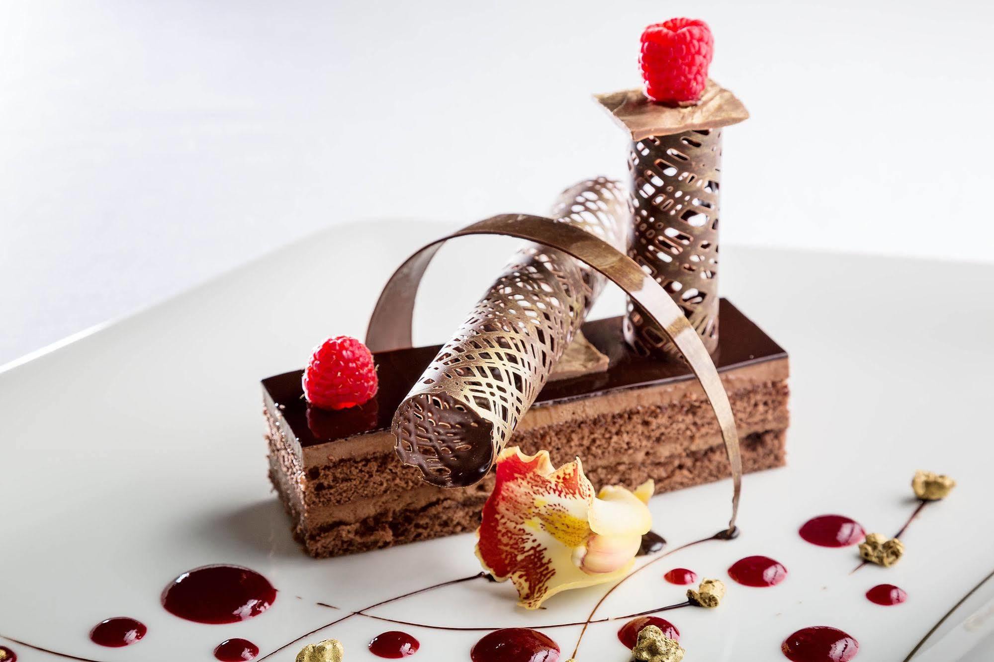 Jabal Omar Hyatt Regency Makkah Hotel Mekka Kültér fotó The photo features an artistic dessert presentation. It includes a rectangular chocolate base layered with a rich chocolate mousse or cake. Atop the cake, there are three decorative elements: two cylindrical structures made of intricate chocolate or 